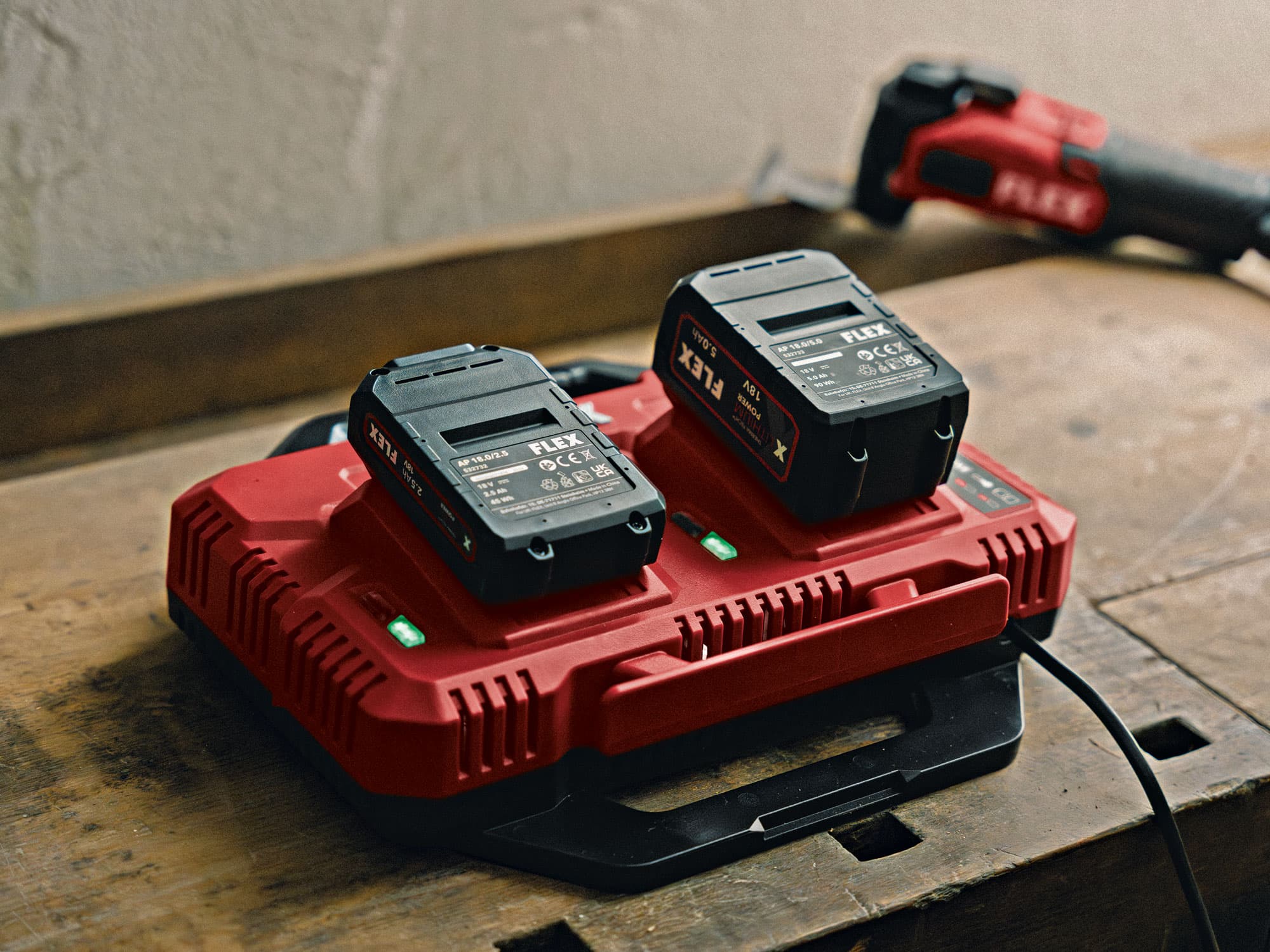 FLEX Doppel SchnellladegerĂ€t mit zwei eingesetzten 18 V Akkus auf einer Werkbank, grĂŒne LED-Anzeige signalisiert aktiven Ladevorgang.