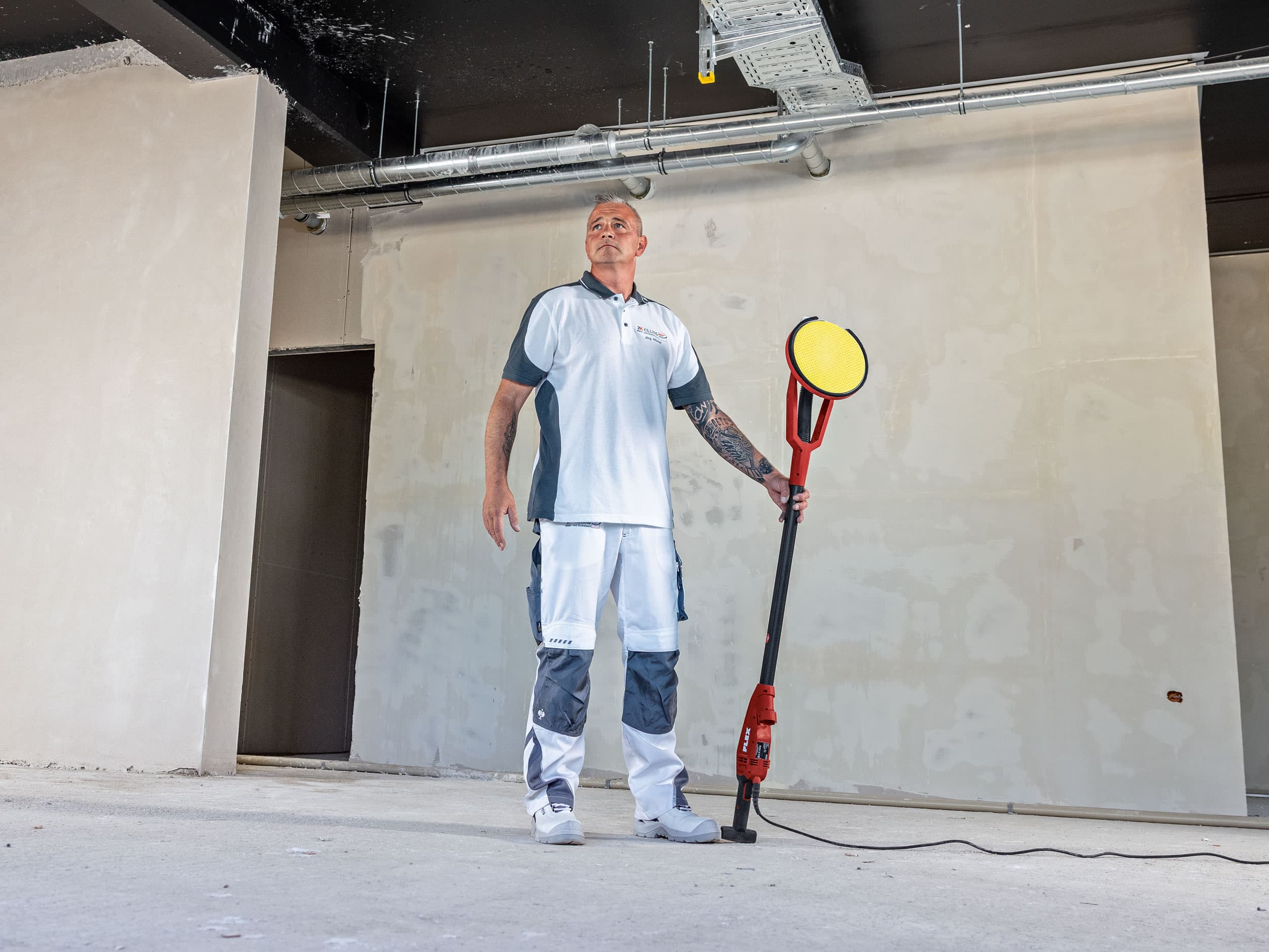 Maler Jörg steht selbstbewusst auf der Baustelle und hält die FLEX GE 6 R – einen Wand- und Deckenschleifer mit rotativem Antrieb – in der Hand.